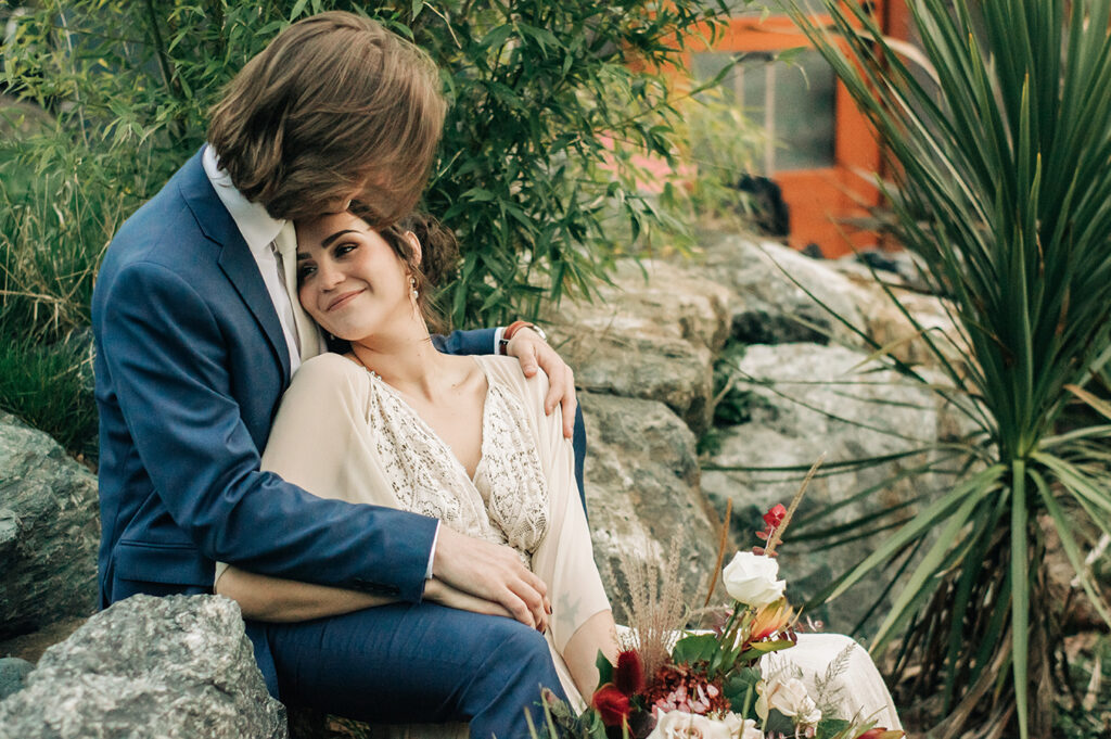 A bride leaning on her groom, seated on rocks surrounded by greenery