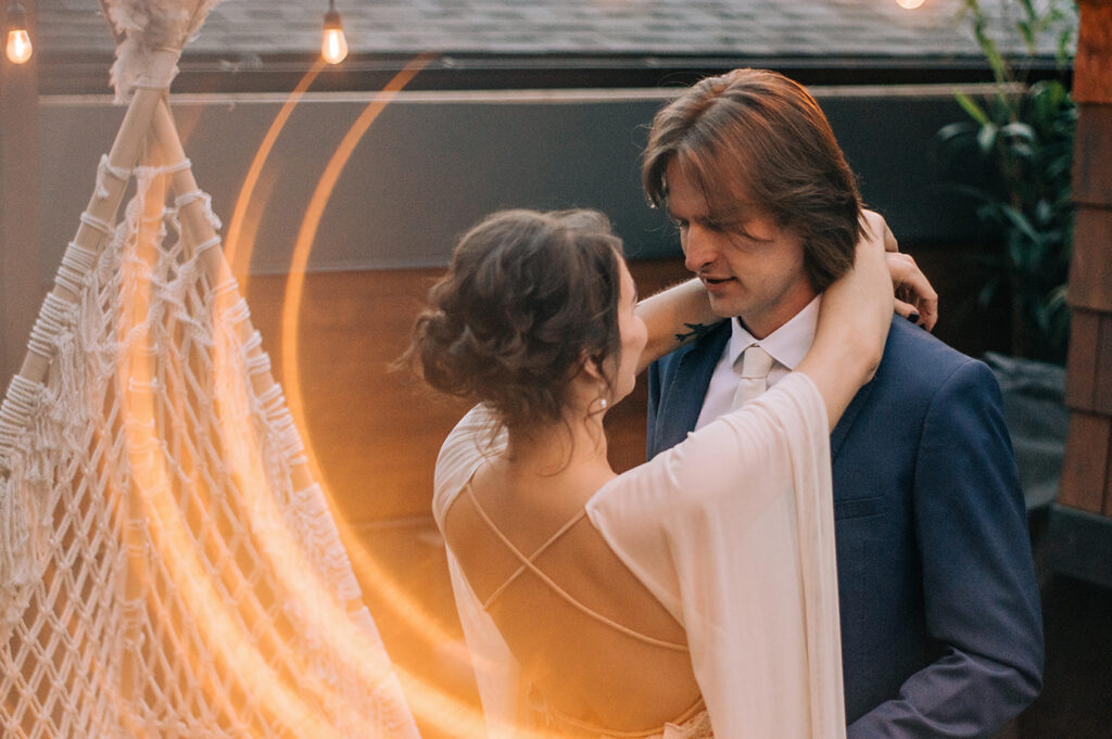 A bride facing her groom with her arms around his neck. it's evening and there is a glow of hanging lights and there's a creative effect around them using the light